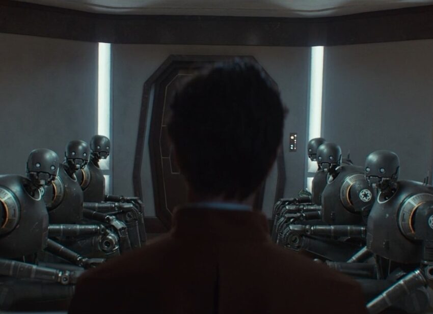 Scene from Andor, Season 2, Episode 8. Syril is pushed into a holding room in which 6 silent KX Security Droids stare blankly at him as he enters. The tension is real.