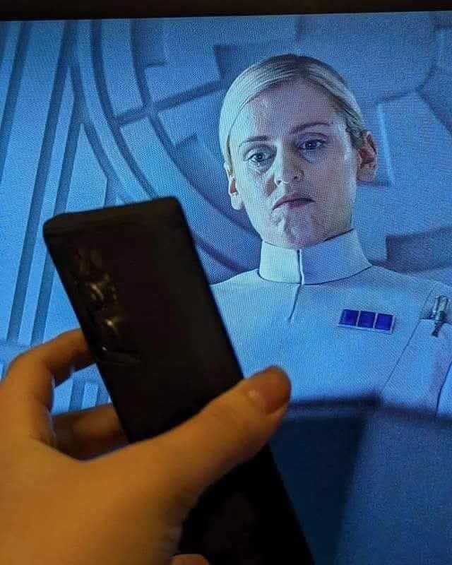 Someone holding a phone screen up a the TV screen as Dedra Meero from Andor looks down into it with her #1 stinkface.