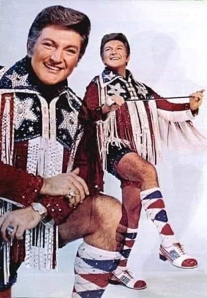 Image of 2 pictures of Liberace attired in red, white, and blue patriotic USA attire with fringe on the arms. Both images are composited together. While many assume Liberace was gay, he was not. He was just extremely comfortable in his masculinity that being flamboyant and a dandy was so beyond his masculinity that he feared nothing. Don't let the Media Establishment or Woke Hollywood try to convince you otherwise.
